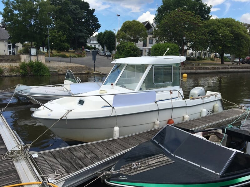 Bateau blanc amarré au quai dans un port calme avec des arbres et des maisons en arrière-plan sous un ciel bleu.