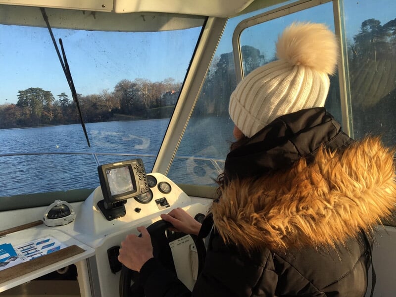 Personne vue de dos portant un bonnet blanc et une veste à capuche avec fourrure, conduisant un bateau sur un lac.