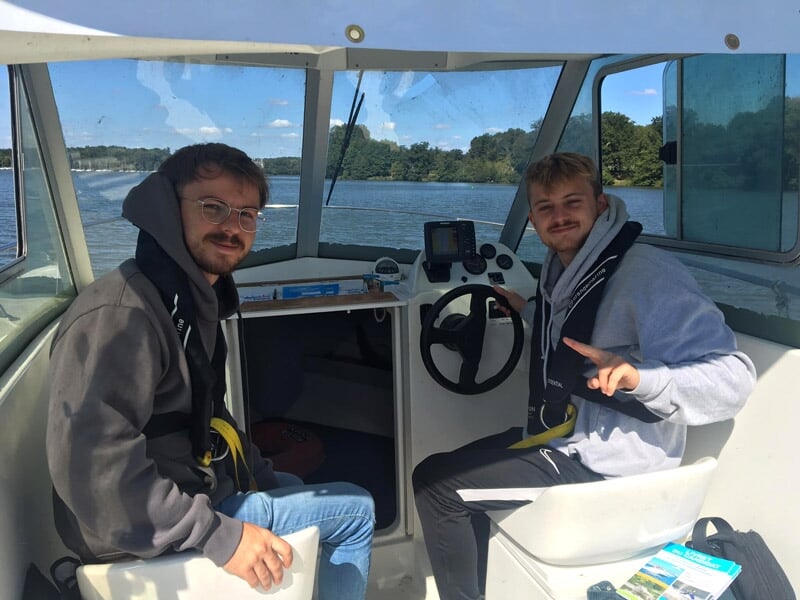 Deux jeunes hommes souriants assis dans un bateau, l'un au volant, avec un paysage d'eau et d'arbres en arrière-plan.