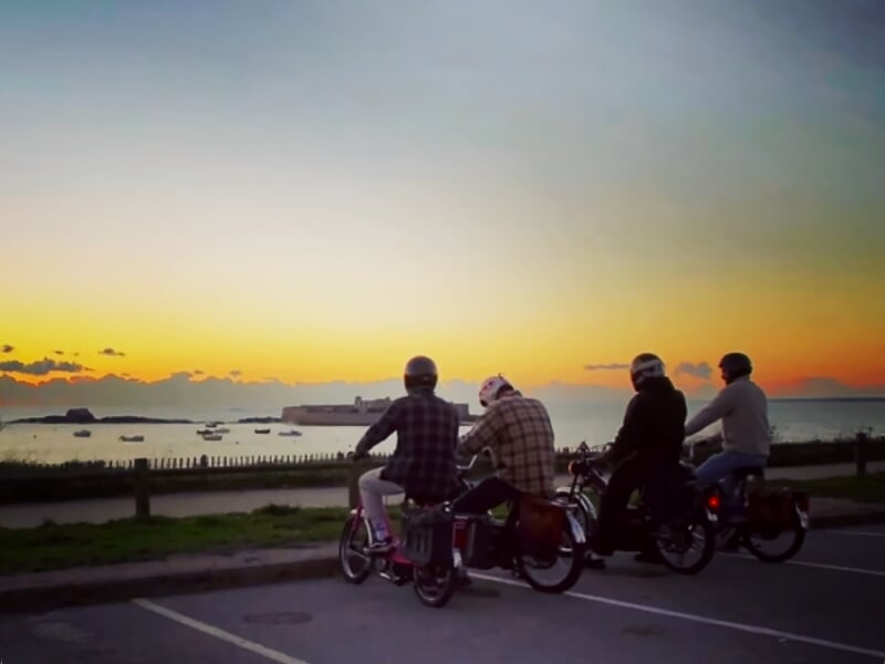 Quatre personnes assises sur des mobylettes face à la mer au coucher du soleil, avec bateaux et digue en arrière-plan.