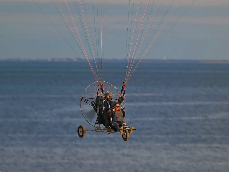 Deux personnes en paramoteur volant au-dessus de la mer avec un ciel clair en arrière-plan.