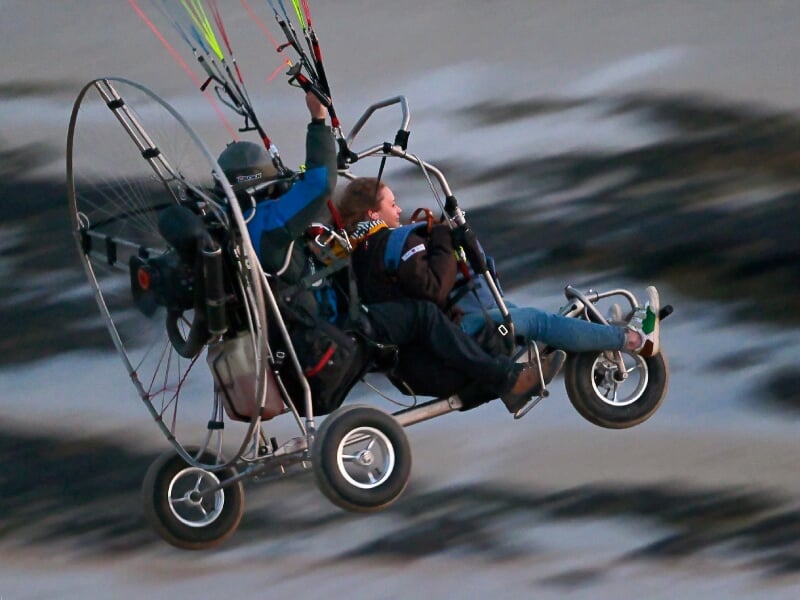 Paramoteur avec deux passagers volant près d'une côte rocheuse, mouvement visible.