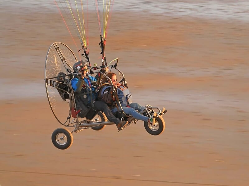 Vol en paramoteur avec deux personnes au-dessus d'une plage au sable doré au coucher du soleil.