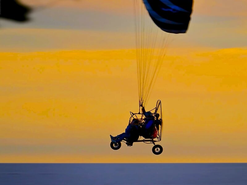 Silhouette d'un paramoteur avec deux passagers volant au coucher du soleil sur un ciel orangé.