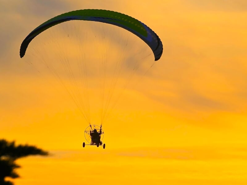 Paramoteur vu de dos avec voile verte et bleue, volant dans un ciel orange au coucher du soleil.