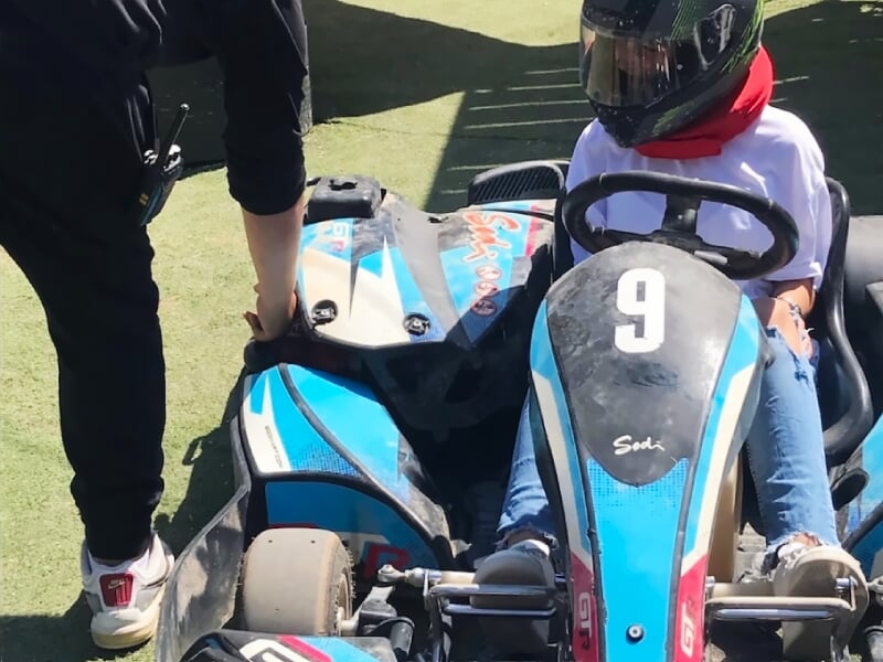 Enfant casqué assis dans un kart bleu numéro 9, prêt à piloter sur une piste extérieure ensoleillée.