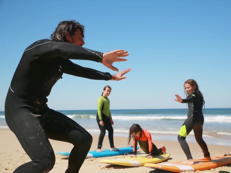 Un instructeur en combinaison noire enseigne le surf à trois enfants sur la plage avec planches colorées et mer calme.