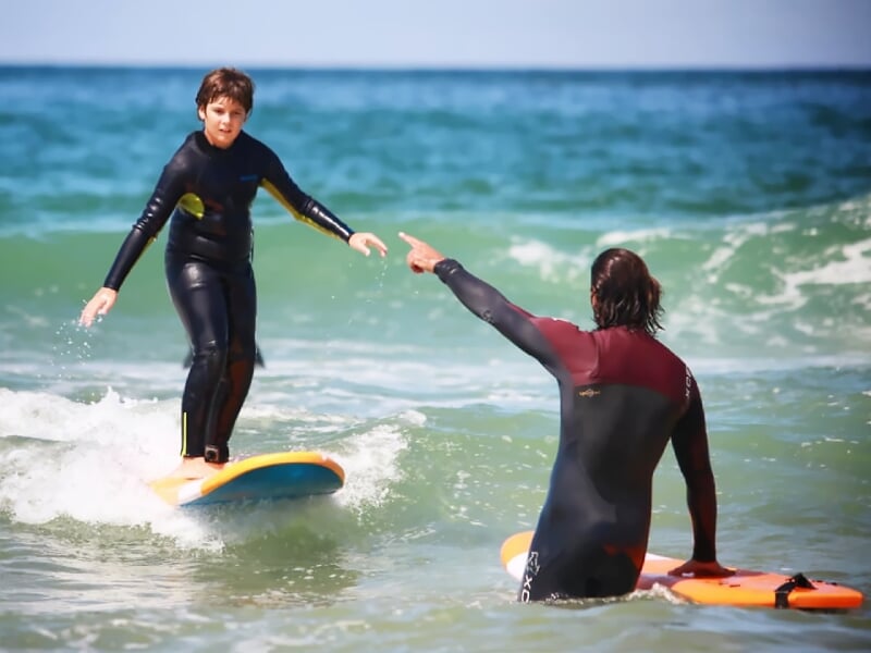 Un enfant en combinaison de surf debout sur une planche, guidé par un instructeur assis dans l'eau qui lui montre la direction.