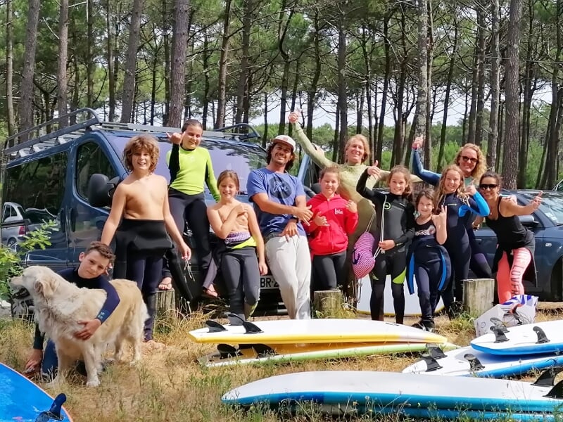 Groupe de jeunes surfeurs et surfeuses en combinaison, posant joyeusement devant un van et des planches de surf en forêt.