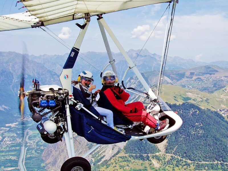 Deux personnes en ULM volant au-dessus des montagnes verdoyantes sous un ciel bleu clair et ensoleillé.
