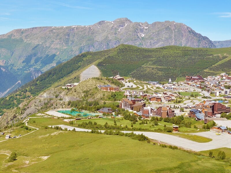 Vue panoramique d'un village de montagne entouré de collines vertes et de routes sinueuses en été.