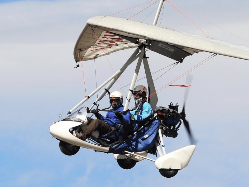 Deux passagers en ULM biplace volant dans un ciel clair, équipés de casques et lunettes de protection.
