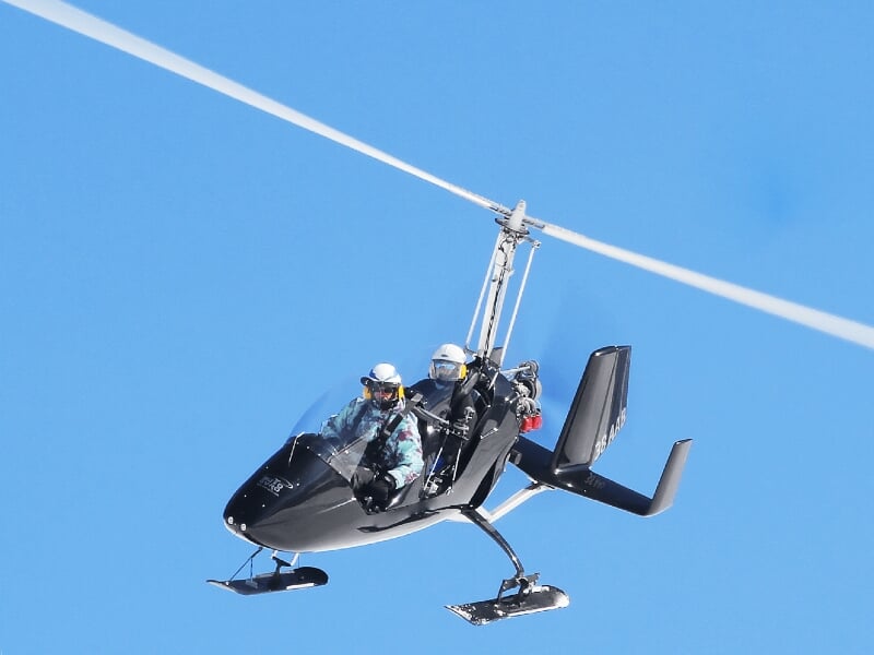 Gyrocoptère noir avec deux passagers en vol dans un ciel bleu dégagé, en pleine activité aérienne.