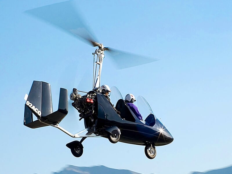 Gyrocoptère noir en vol avec deux personnes à bord, sur fond de ciel bleu clair et montagnes au loin.