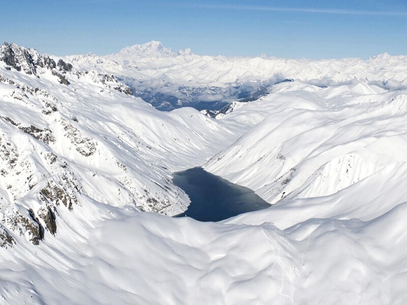 Vue aérienne d'une vallée enneigée avec un lac gelé entouré de montagnes blanches sous un ciel clair.