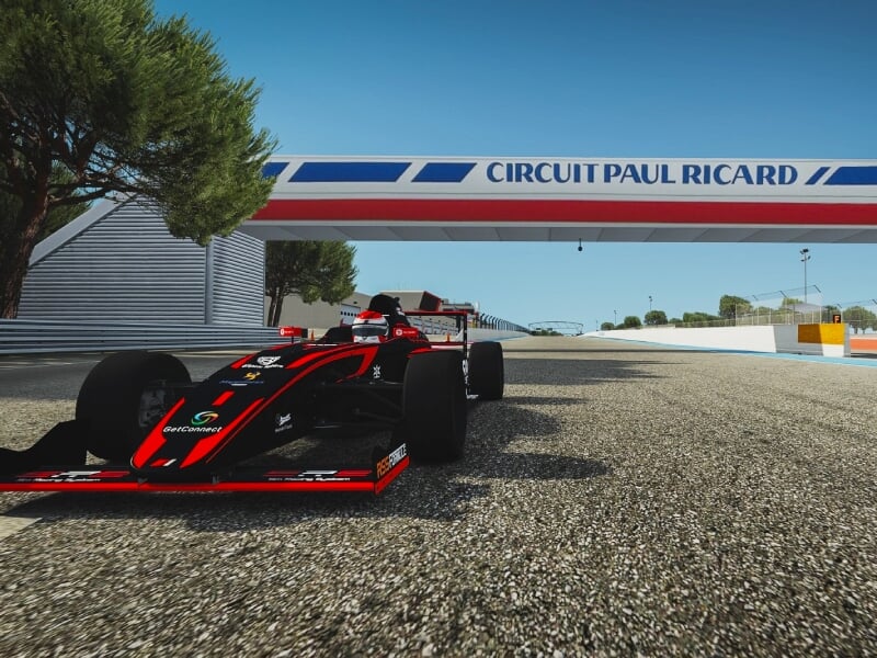 Voiture de course noire et rouge sur le circuit Paul Ricard, passant sous une arche marquée du nom du circuit, avec des arbres et un ciel bleu, soulignant l'importance de ce lieu pour les courses auto