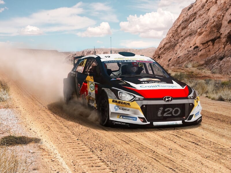 Voiture de rallye Hyundai i20, portant des sponsors tels que Red Bull et Bardahl, dérapant sur une piste poussiéreuse en pleine nature, avec des montagnes en arrière-plan, illustrant la vitesse et la 