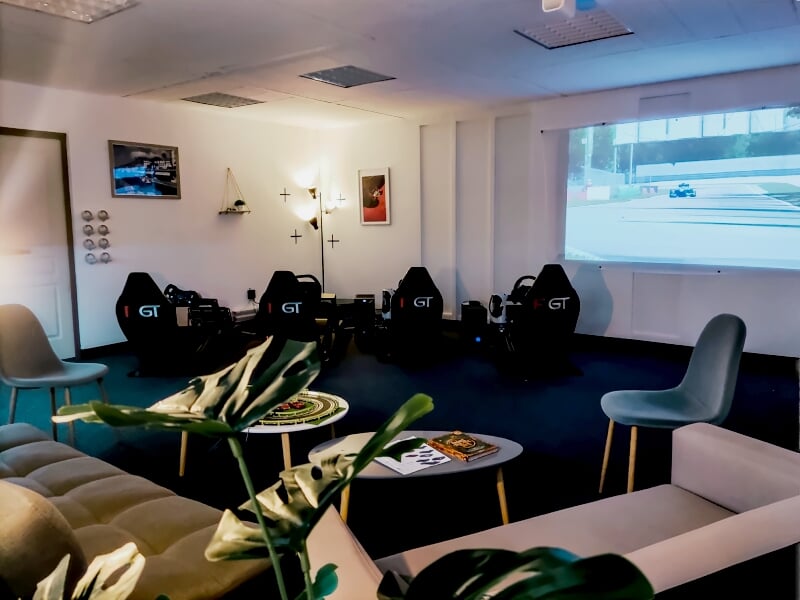 Salle de simulation de course équipée de plusieurs simulateurs GT, situés devant un grand écran projetant une course automobile. La pièce dispose également d'un coin détente avec des canapés, des chai