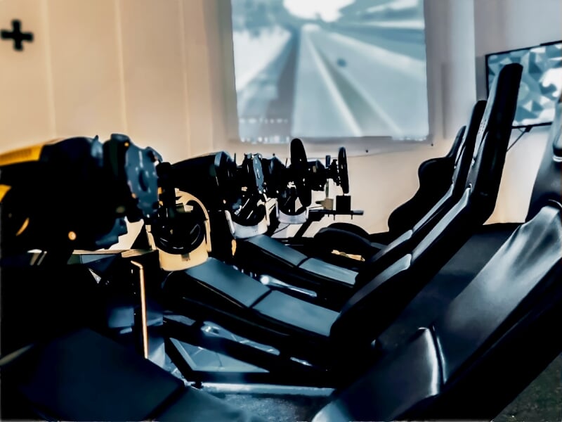 Plusieurs simulateurs de course alignés, équipés de volants et de sièges baquets noirs, avec un écran projetant une vue d'une piste de course à l'arrière-plan, créant une ambiance immersive pour les p