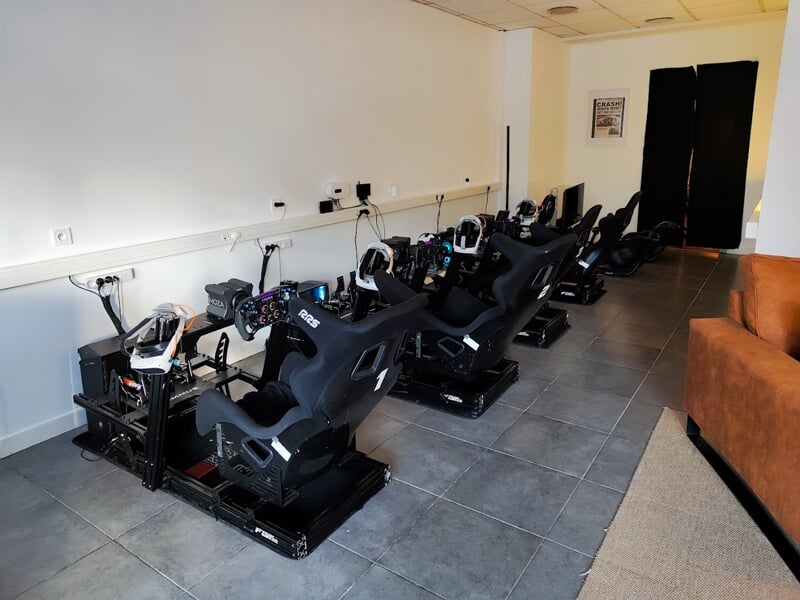 Salle équipée de simulateurs de course alignés contre un mur blanc, avec sièges baquets noirs RSeat, volants racing, pédaliers haut de gamme et écrans individuels. Sol en carrelage gris et câblage app