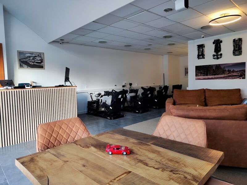 Vue d’ensemble d’un espace de simulation avec cinq simulateurs de course, un comptoir d’accueil en bois clair, un canapé marron, une table en bois massif et des décorations murales sur le thème de l’a