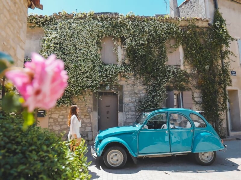 Voiture bleue Citroën 2CV garée devant une maison en pierre couverte de lierre avec une fleur rose au premier plan.