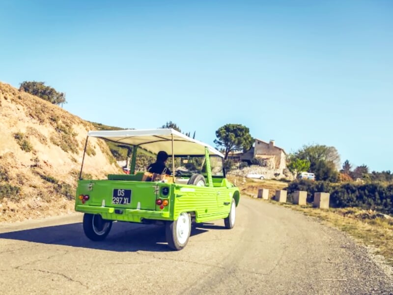 Voiture verte vintage avec toit ouvert roulant sur une route de campagne avec une maison en arrière-plan.