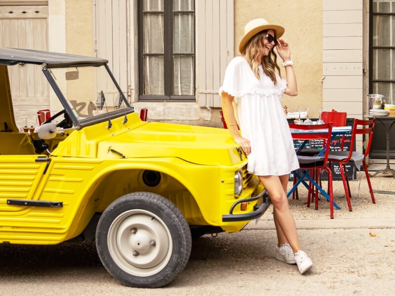 Femme en robe blanche et chapeau posant à côté d'une voiture jaune vintage devant un café en plein air.