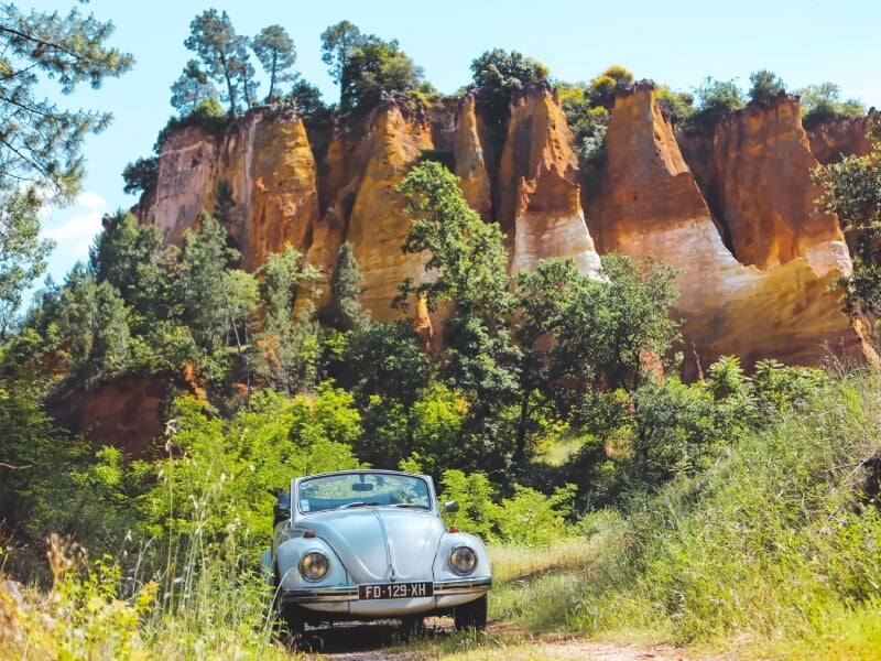 Voiture décapotable blanche garée devant des falaises rouges et jaunes dans un paysage naturel ensoleillé.
