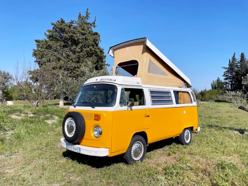 Combi orange avec toit relevé garé sur une pelouse verte avec des arbres et un ciel bleu en arrière-plan.