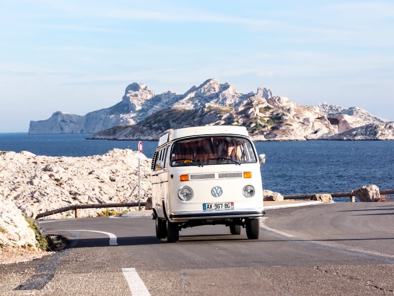 Combi blanc roulant sur une route côtière avec vue sur la mer et des falaises rocheuses au loin.