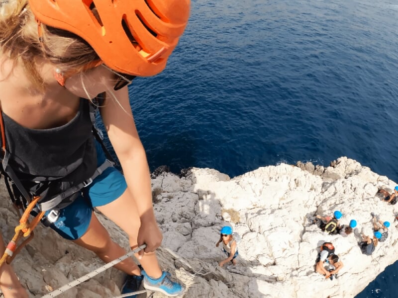 Personne équipée d'un casque orange escaladant une falaise rocheuse au-dessus de la mer bleue profonde.