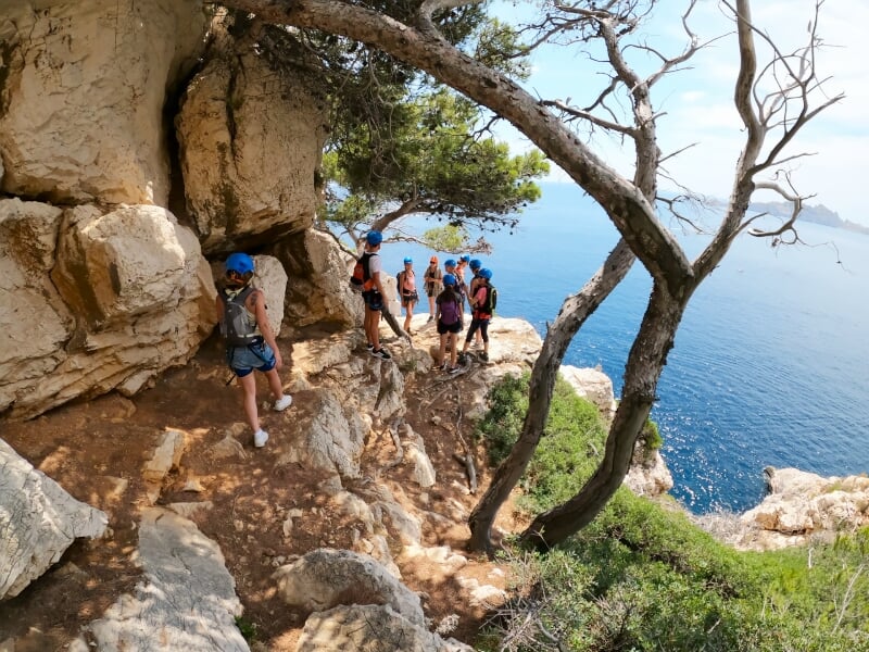 Groupe de grimpeurs avec casques bleus marchant sur un sentier rocheux en bord de mer sous un ciel clair.