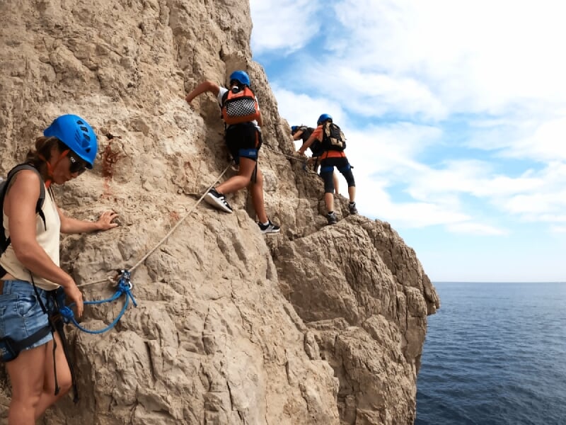 Trois grimpeurs avec casques bleus progressant sur une paroi rocheuse verticale au bord de la mer.