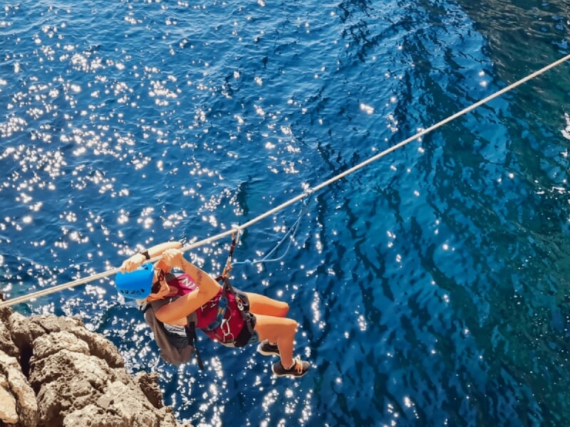 Personne en casque bleu suspendue à une tyrolienne au-dessus d'une mer scintillante sous un ciel ensoleillé.