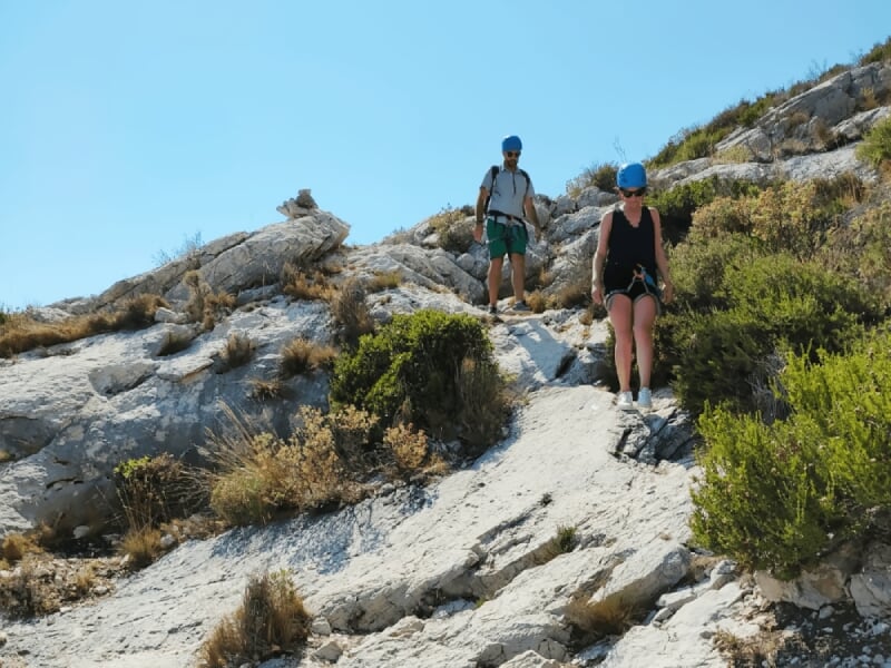 Deux personnes avec casques bleus descendant prudemment une pente rocheuse en terrain naturel ensoleillé.