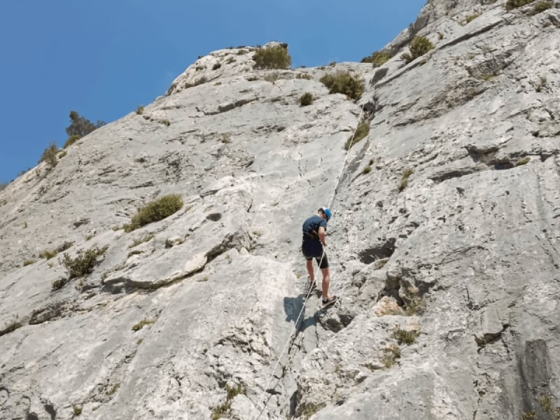 Grimpeur en casque bleu escaladant une falaise rocheuse abrupte sous un ciel partiellement nuageux.