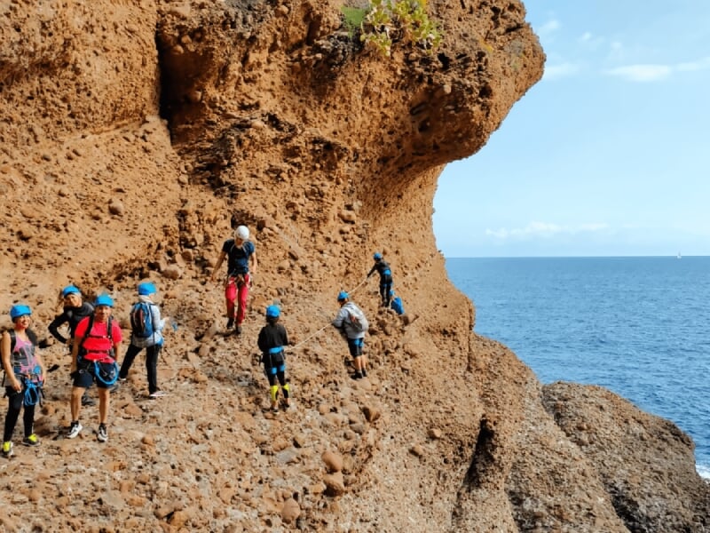 Groupe de personnes équipées de casques et harnais marchant sur un sentier rocheux au bord de la mer.
