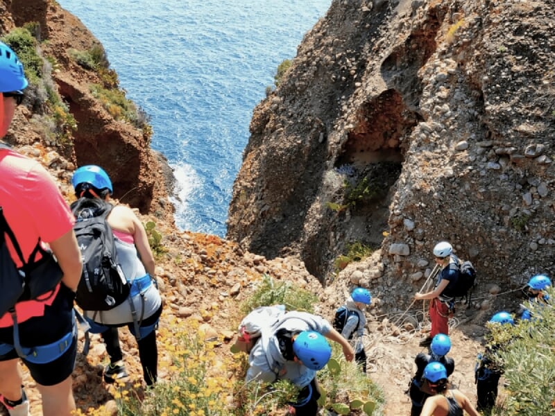 Groupe de grimpeurs avec casques bleus descendant un sentier rocheux escarpé en bord de mer.