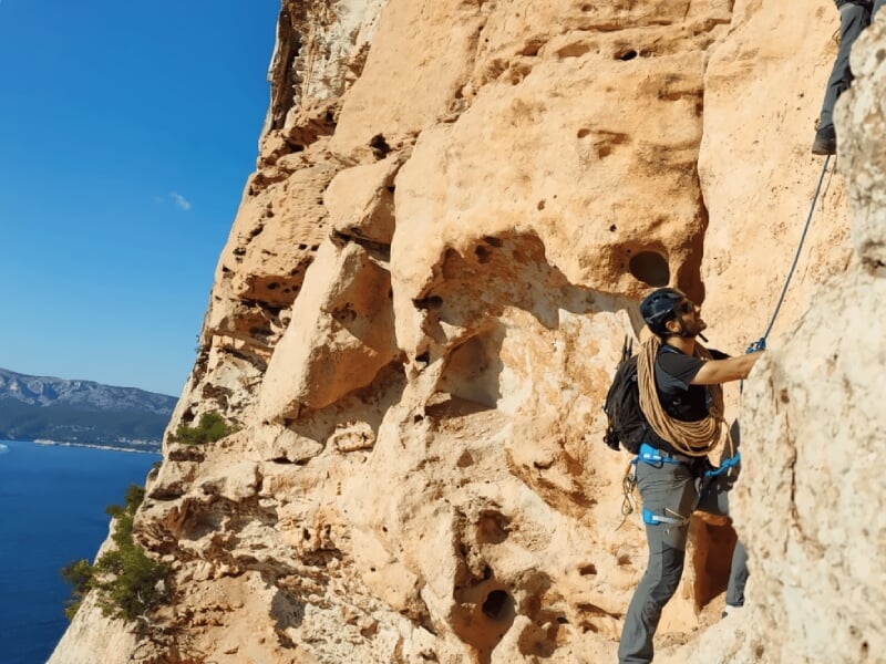 Homme en casque noir escaladant une falaise rougeâtre avec la mer visible en arrière-plan.