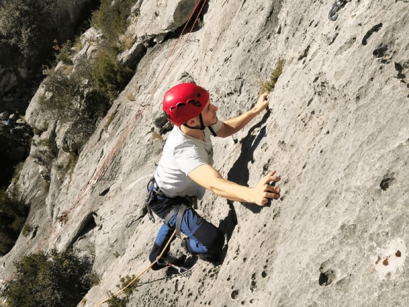 Grimpeur en casque blanc aidant une autre personne à escalader une paroi rocheuse ensoleillée.