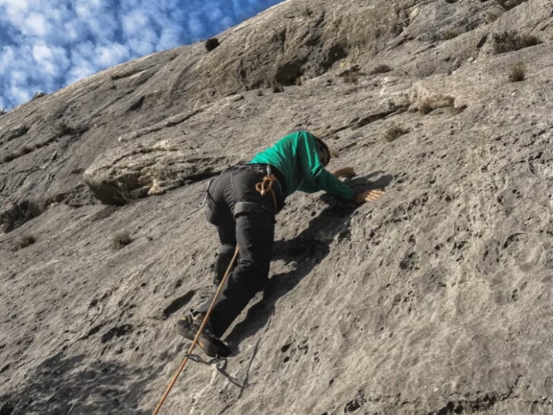 Escalade en extérieur, grimpeur sur paroi rocheuse grise, ciel partiellement nuageux, corde de sécurité fixée.