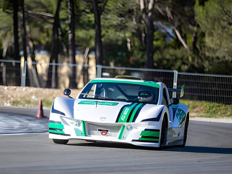 Voiture de course blanche et verte sur circuit, en virage, avec arbres en arrière-plan.