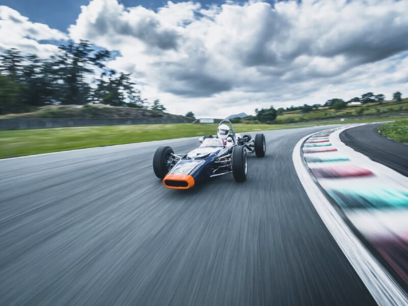 Voiture de course noire et orange prise en photo en mouvement sur un circuit avec bordure colorée, pilote en casque, ciel nuageux