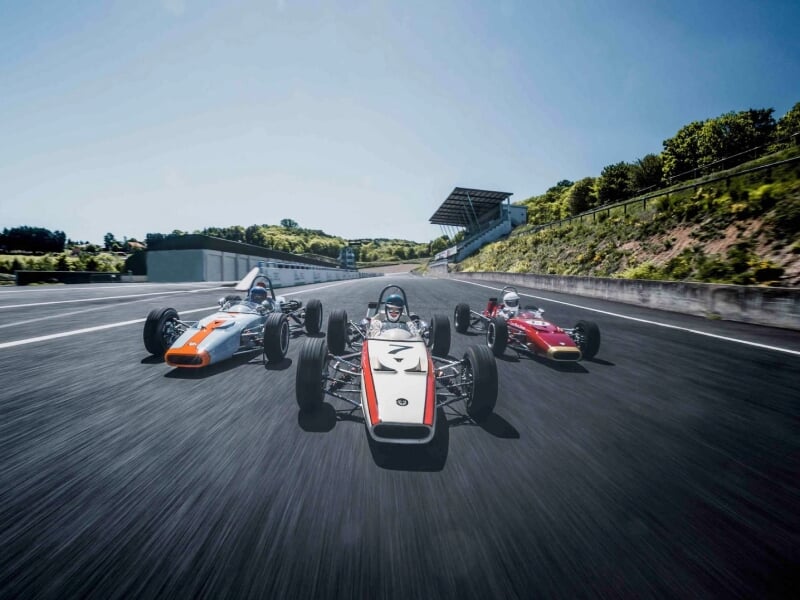 Trois voitures de course anciennes roulant côte à côte sur une piste, pilote en casque visible dans chaque voiture, fond de verdure et ciel dégagé