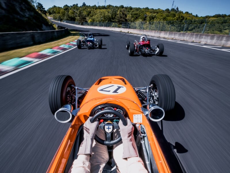 Vue depuis l'intérieur d'une voiture de course orange sur un circuit, pilote portant des gants blancs tenant le volant, deux autres voitures de course visibles devant