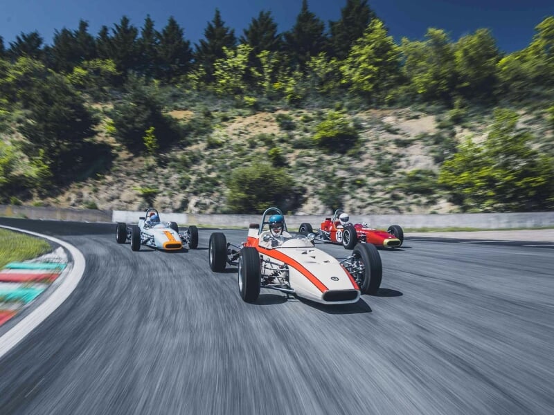 Trois voitures de course anciennes en action sur un circuit, pilote avec casque visible dans chaque voiture, environnement verdoyant et ciel bleu