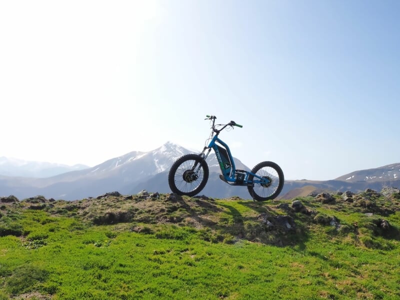 Trottinette électrique bleue posée sur un terrain herbeux avec montagnes enneigées en arrière-plan.