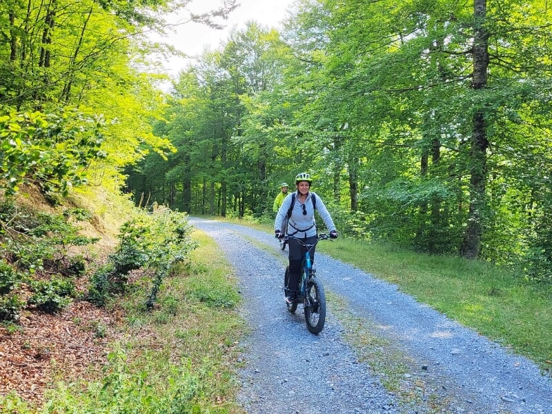 Homme en casque roulant sur une trottinette électrique sur un chemin forestier entouré d'arbres verts.