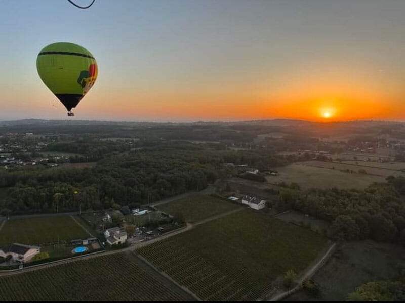 Montgolfière verte flottant au-dessus d'un paysage rural au coucher du soleil, illuminant les champs et les maisons en contrebas